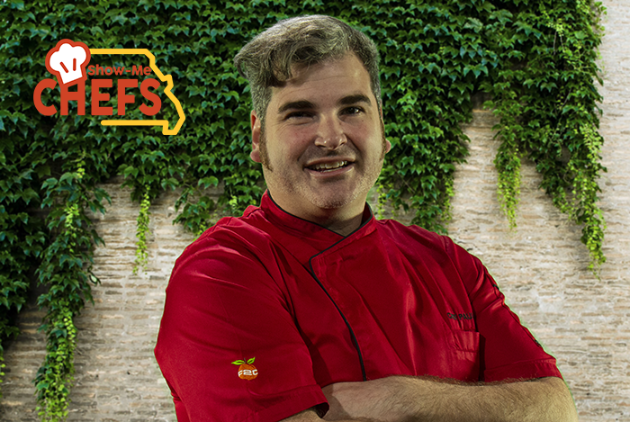 Chef Paul Allen grins in his red chef coat in front of a brick wall covered in ivy